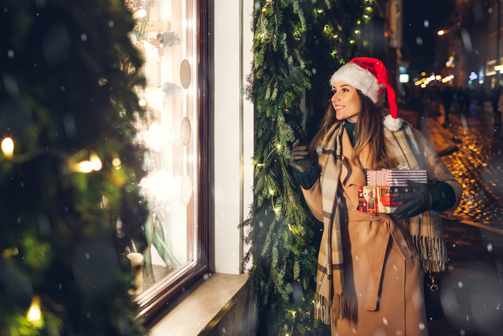 Happy woman in Santa's hat holding presents gift boxes by store showcase on city street at Christmas season. Girl walking by decorated festive shops under falling snow