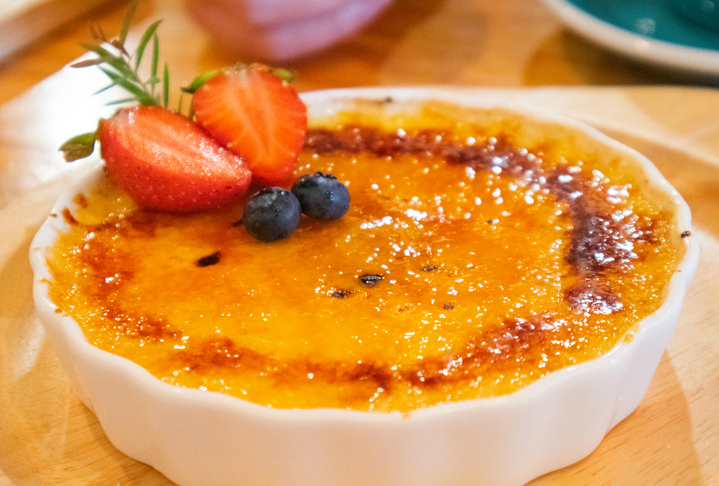 A golden yellow Creme Brulee topped with crunchy brown sugar topped with strawberries and blueberries to be included in a white glass plate on the dining table after eating.