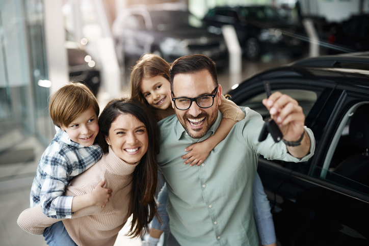Happy parents piggybacking their small kids after receiving keys for their new car in a showroom.