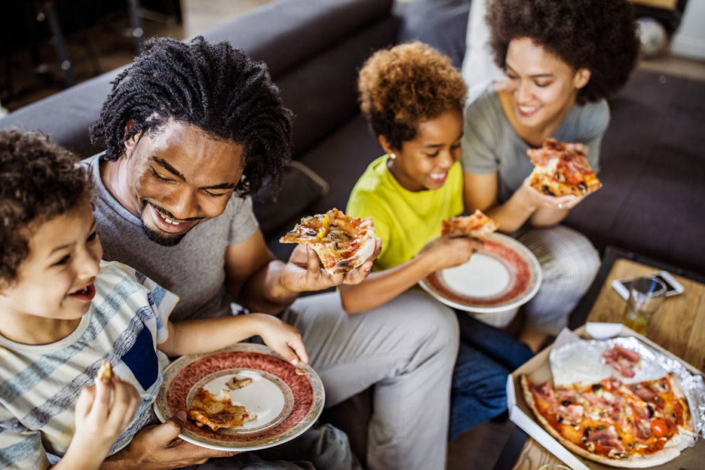 Happy black parents and their small kids eating pizza for lunch in the living room.