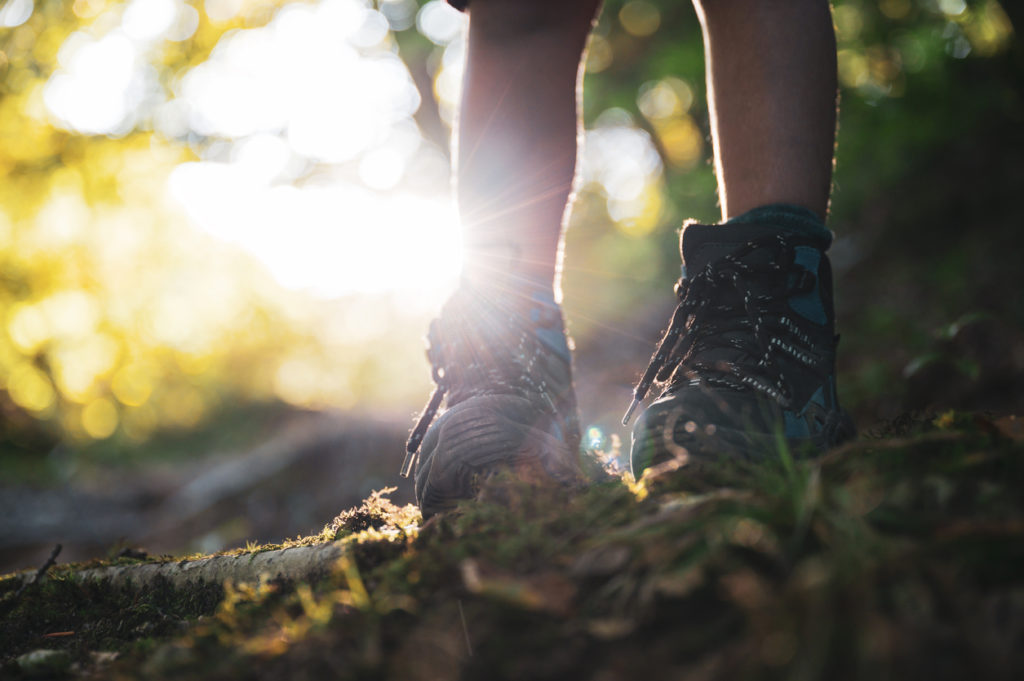 Low angle view of a child's legs in hiking boots standing on mountain trail back lit by the glowing sun.