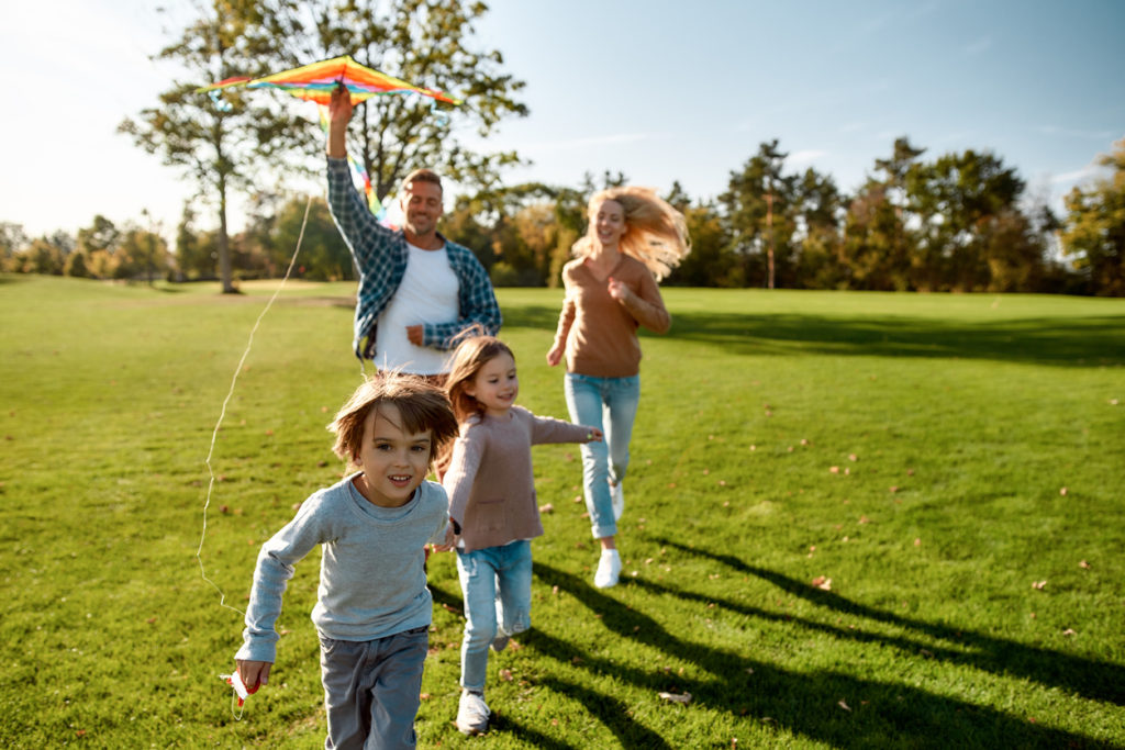 Portrait of cheerful parents with two kids running with kite in the park on a sunny day.